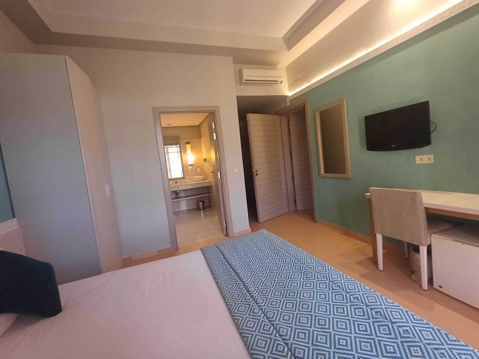 A bedroom with a bed, a desk, a chair, a TV, and a bathroom visible through an open door.