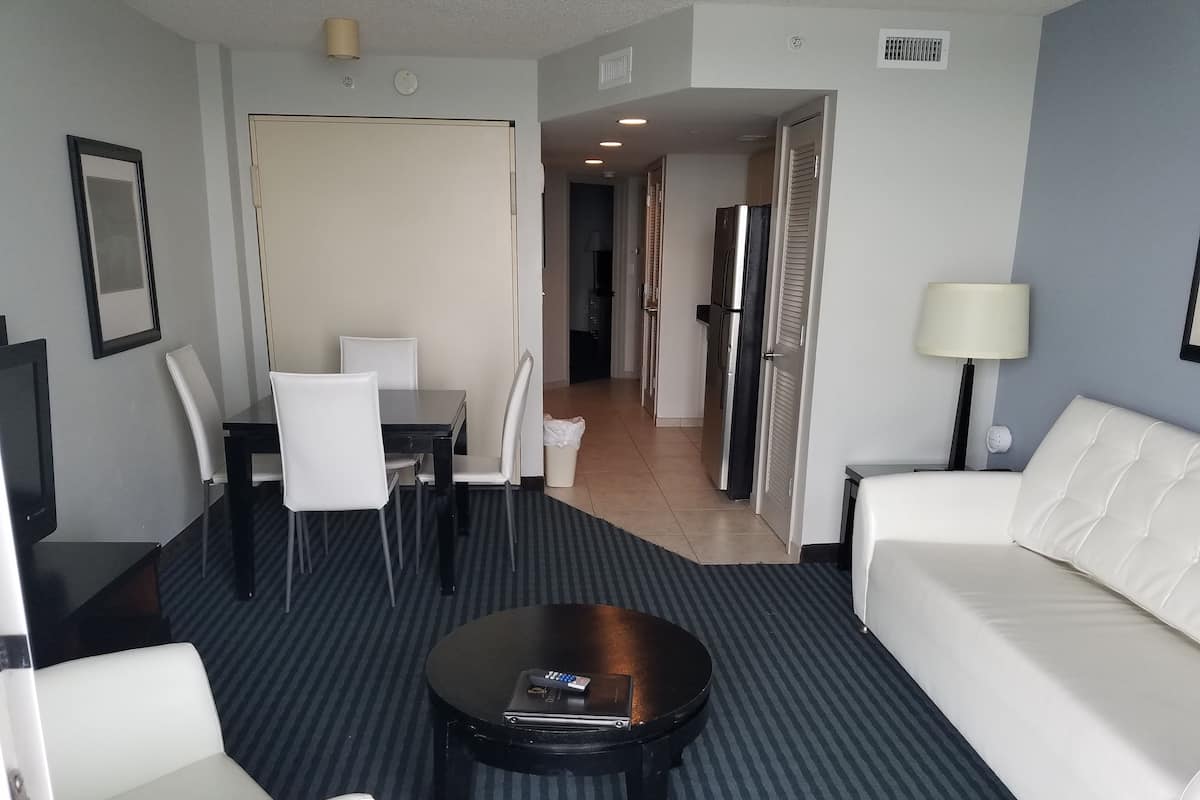 A hotel room with a sofa, a dining table with chairs, a coffee table, a TV, and a lamp.