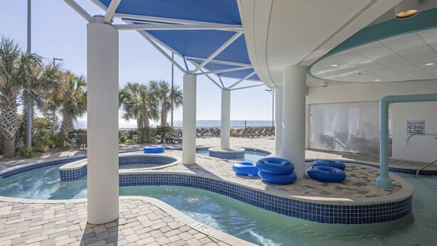 A pool area with multiple hot tubs and lounge chairs, surrounded by palm trees and a view of the ocean. - Oceans One Resort (Myrtle Beach)