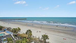 A beach with a pier, pool area, and palm trees. - Oceans One Resort (Myrtle Beach)