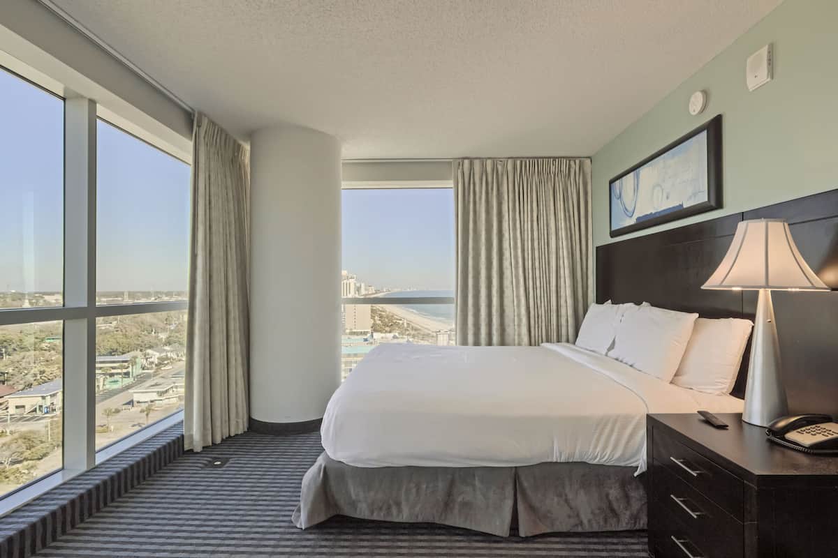 A hotel room with a large bed, a nightstand with a lamp, a phone, and a view of the city and water through large windows.