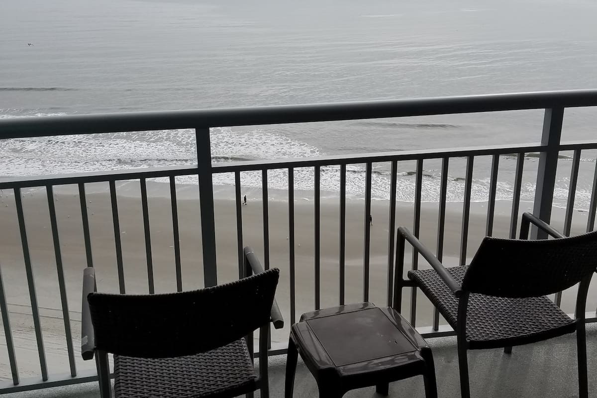 A balcony with two wicker chairs and a small table, overlooking a beach with gentle waves.