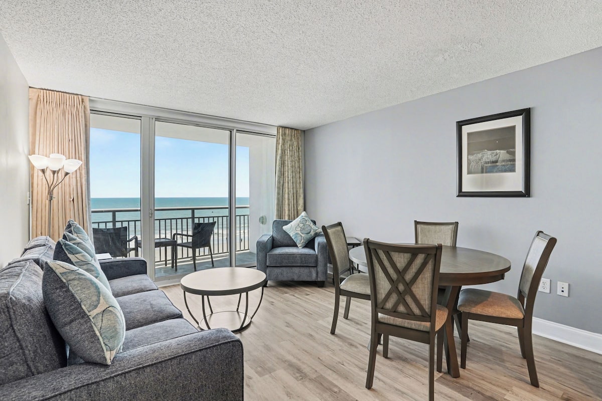 A modern living room with a sofa, chairs, a dining table, a balcony with a view of the sea, and a framed picture on the wall.