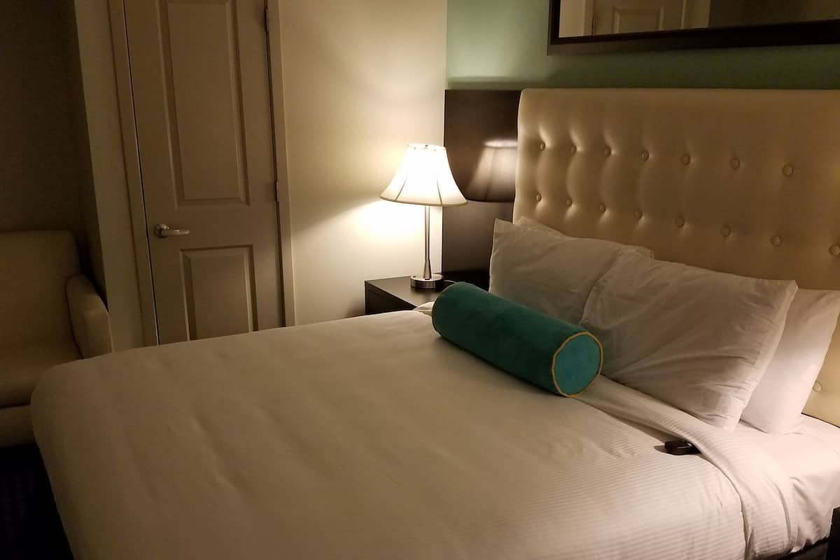 A neatly made bed with a teal pillow, a bedside lamp, and a chair in a hotel room.