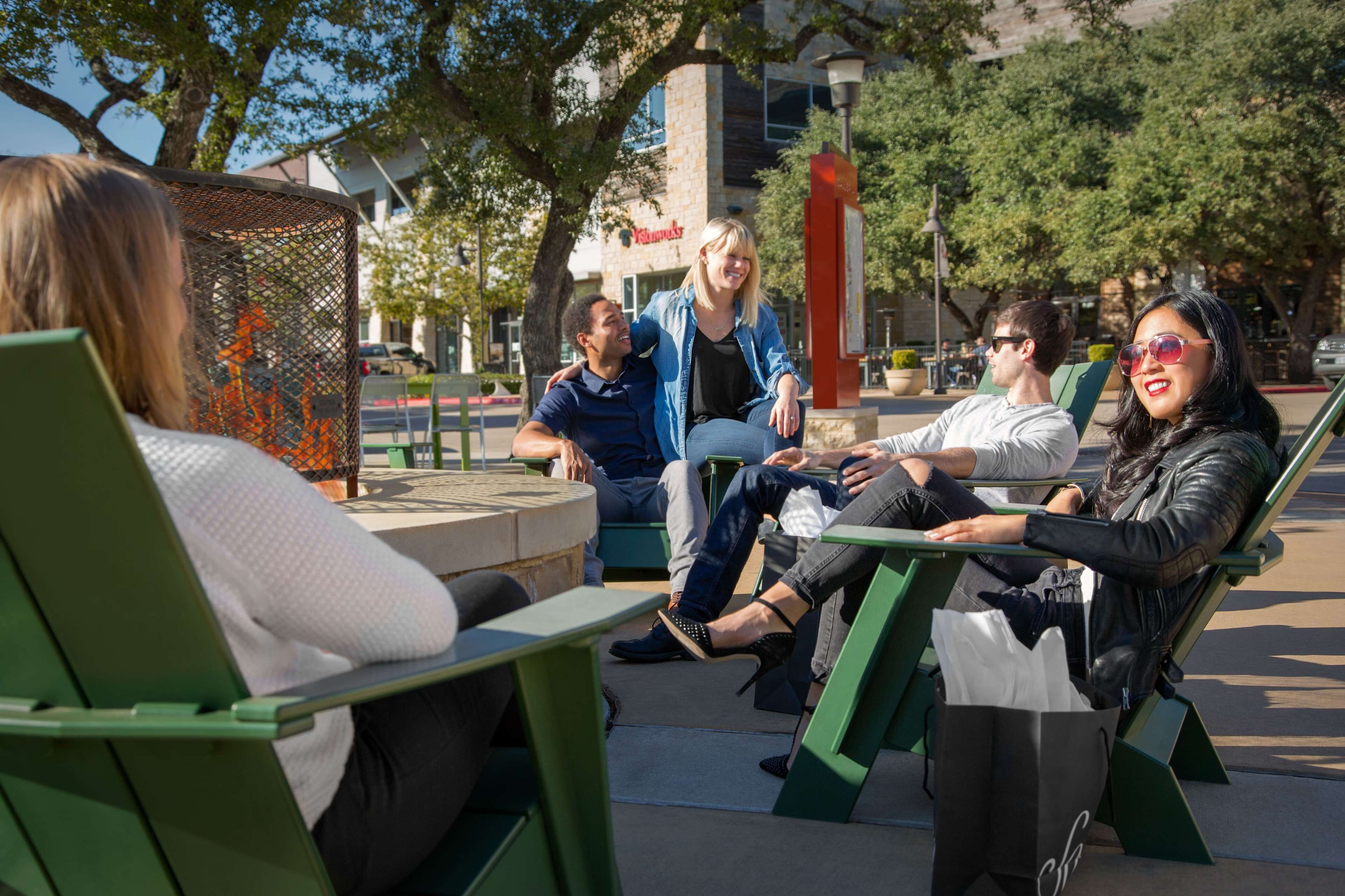 A group of people sitting on green benches in an outdoor setting with trees and a building in the background.