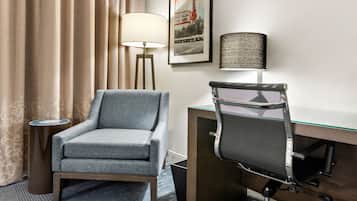 Et hotelværelse med en grå lænestol, et skrivebord med en netvævstol, en lampe og et billede i ramme på væggen.