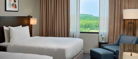 A hotel room with two beds, a desk, a chair, and a large window with a view of greenery.