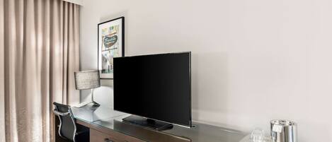 A hotel room with a glass desk, a flat-screen TV, a lamp, a coffee maker, and a framed picture on the wall.