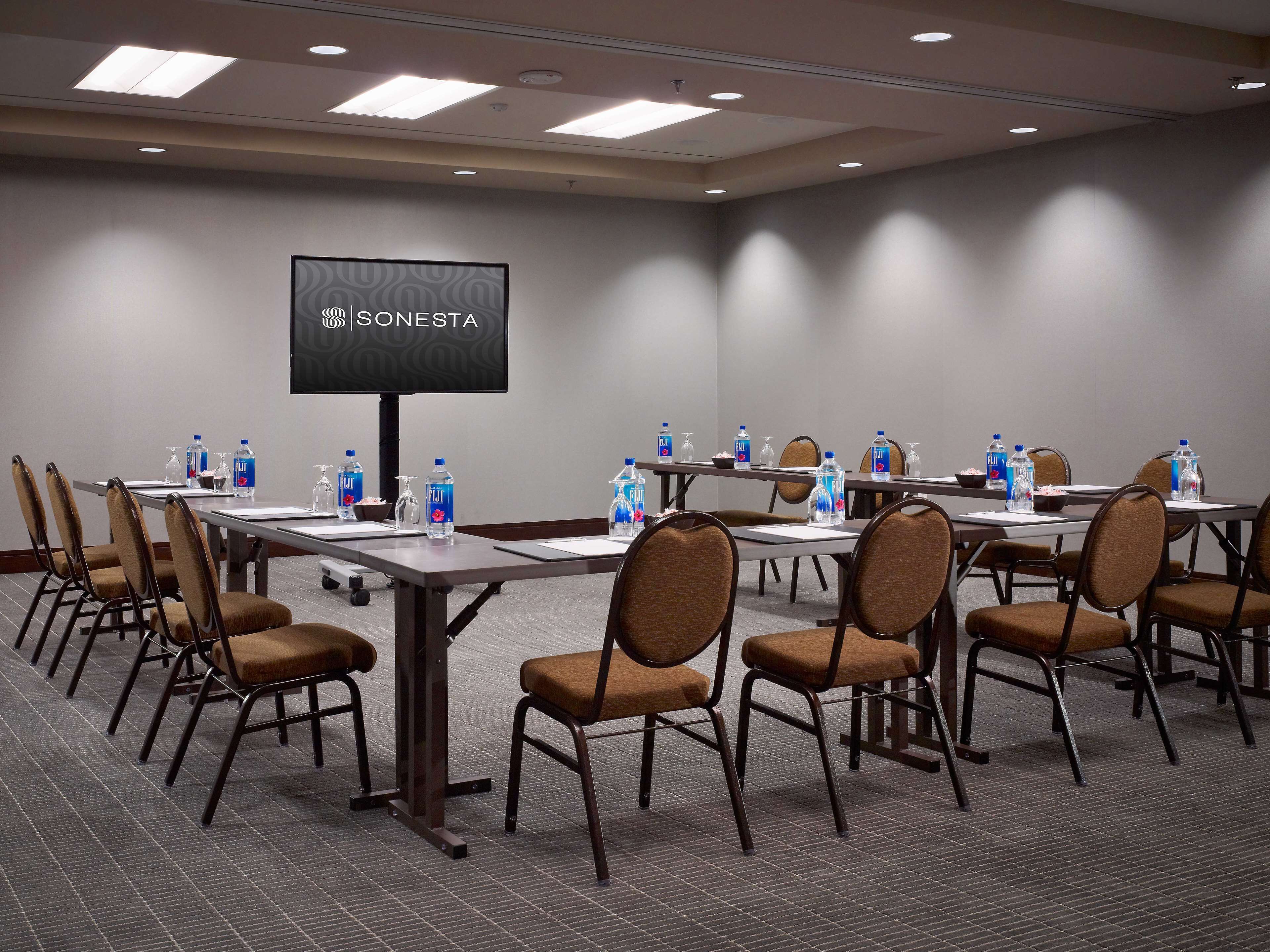 A conference room with a large screen displaying "SONESTA", equipped with water bottles and chairs arranged around tables.