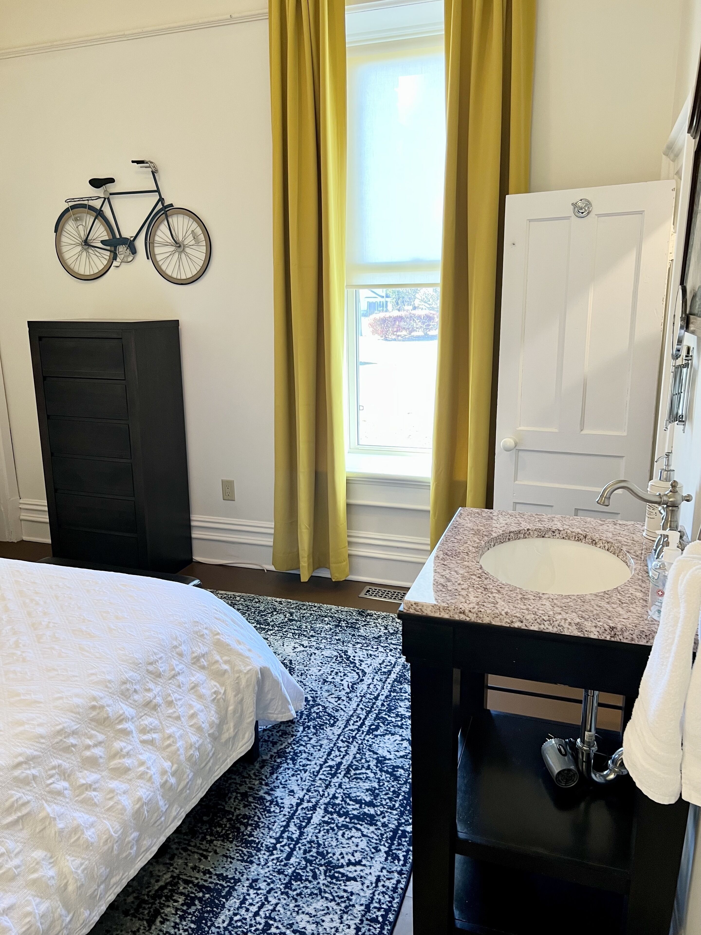 A bedroom with a bed, a dresser, a bathroom with a sink and mirror, a bicycle mounted on the wall, and a window with yellow curtains.