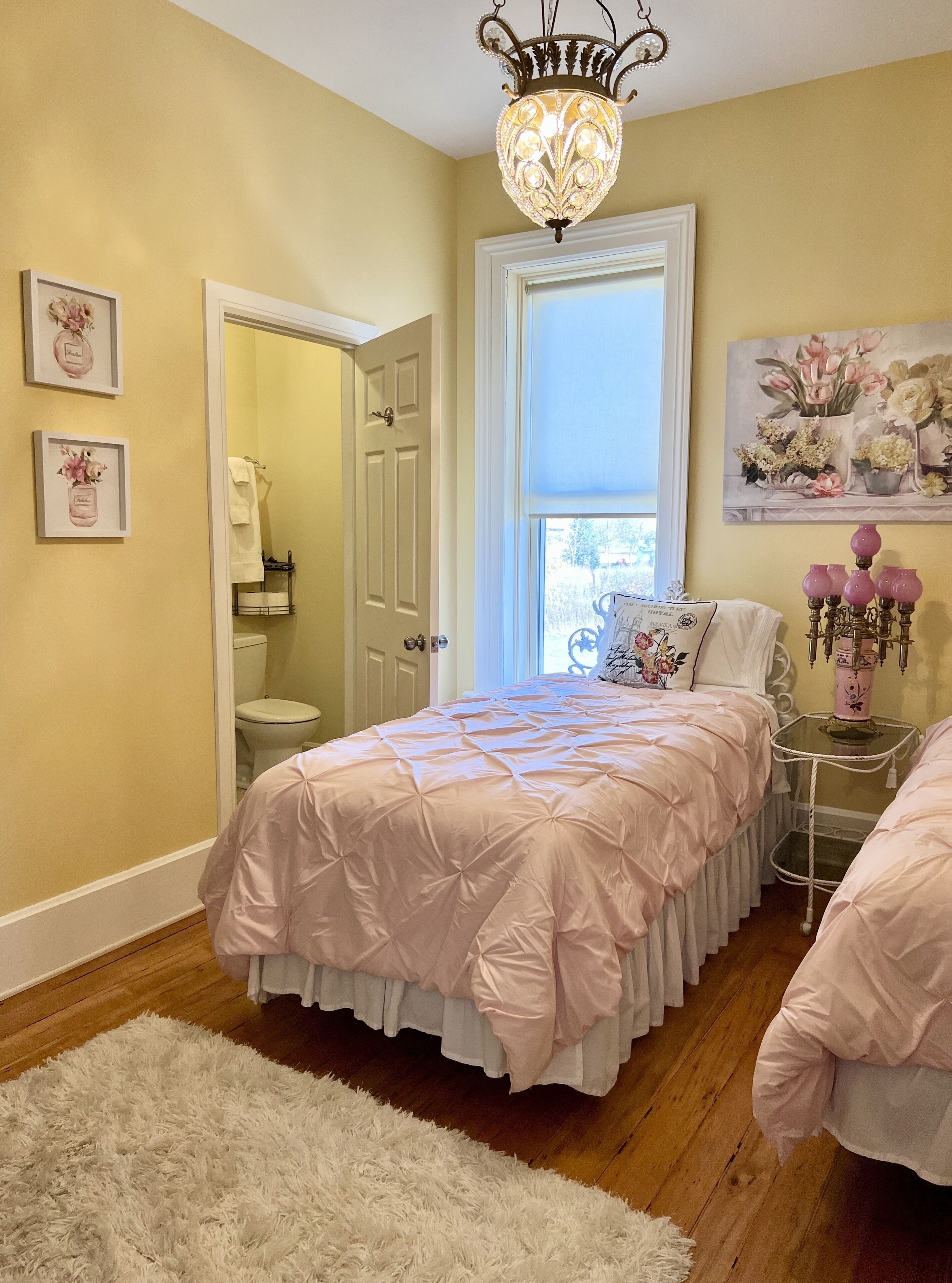 A bedroom with a large bed, a white door leading to a bathroom, a chandelier, and a window with a view.