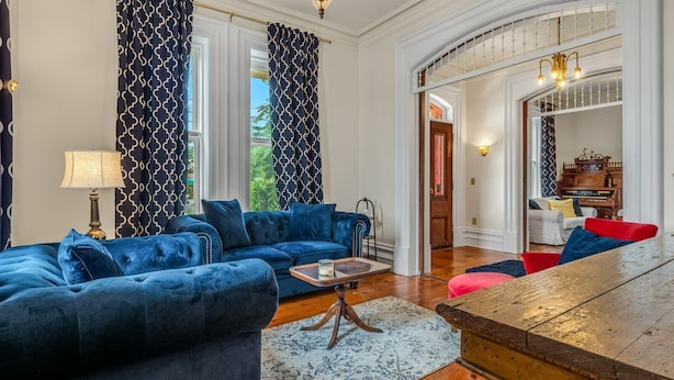 A spacious living room with a blue sofa, a wooden coffee table, and a red chair. There is a lamp, a patterned rug, and a view of the outdoors through large windows. - The Woodbridge House (Kingsville)
