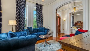 A spacious living room with a blue sofa, a wooden coffee table, and a red chair. There is a lamp, a patterned rug, and a view of the outdoors through large windows. - The Woodbridge House (Kingsville)