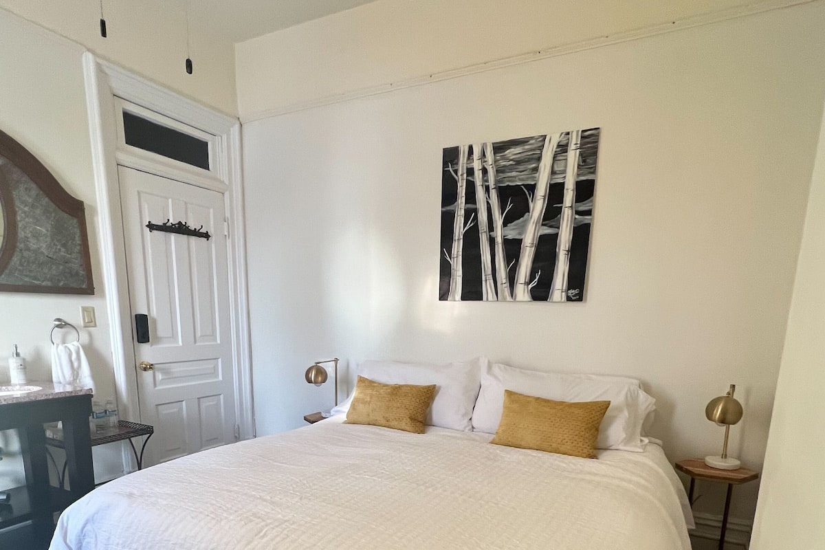 Un dormitorio con una cama grande, una mesita de noche con lámpara, una pequeña mesa con lámpara, una puerta y un cuadro en la pared.