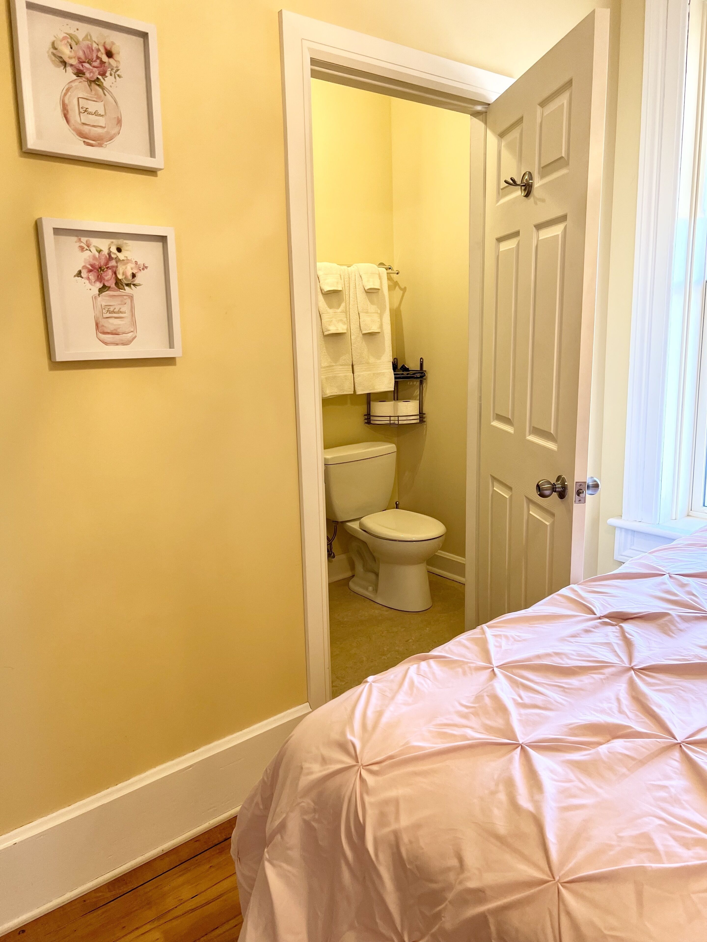 Un dormitorio con una cama, una puerta que da a un baño y dos cuadros con motivos florales en la pared.