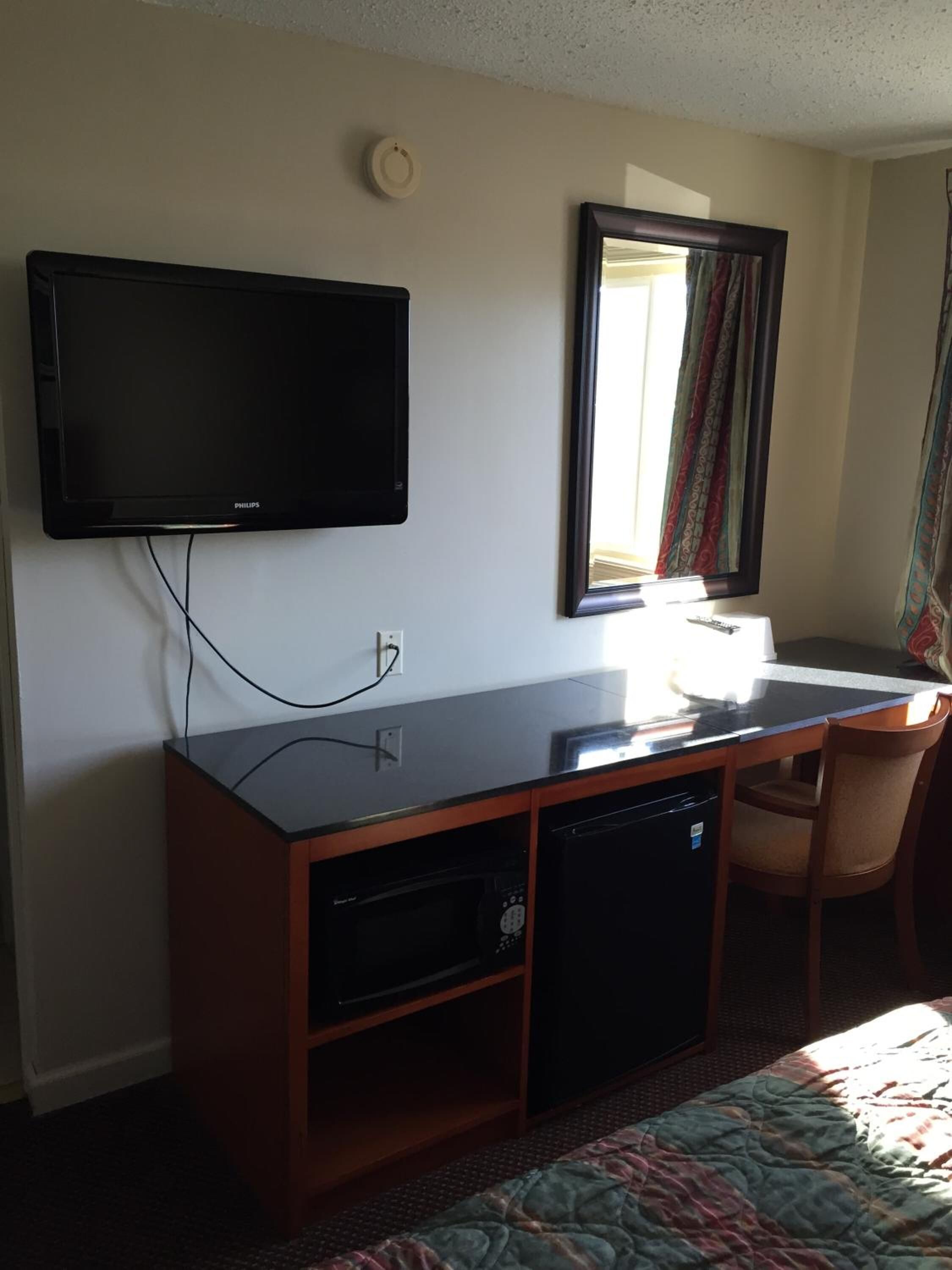 Habitación de hotel con televisor de pantalla plana montado en la pared, un escritorio pequeño con una silla, microondas y lavavajillas.