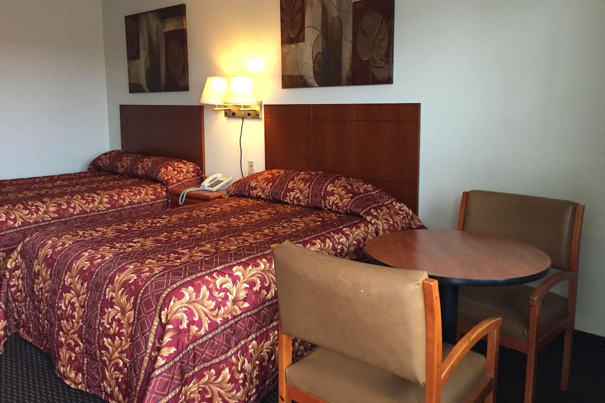 A hotel room with two beds, a desk, and a chair. There is a lamp on the desk and a phone on one of the beds.