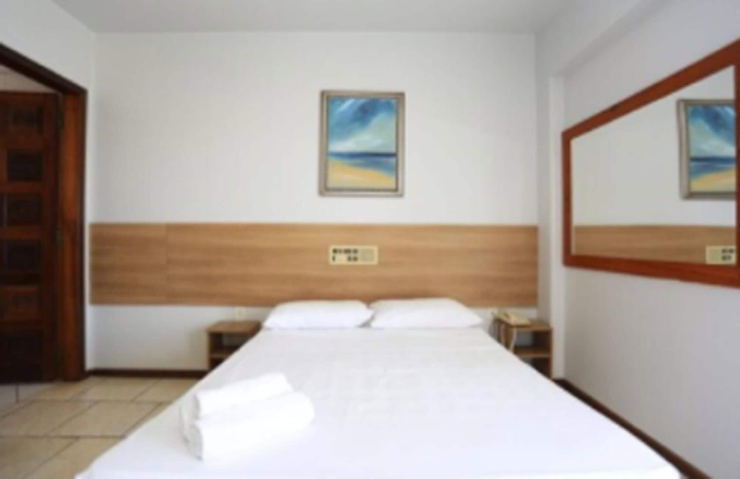 A hotel room with a wooden headboard, a bed with white linens, a nightstand, and a painting of a beach scene on the wall.