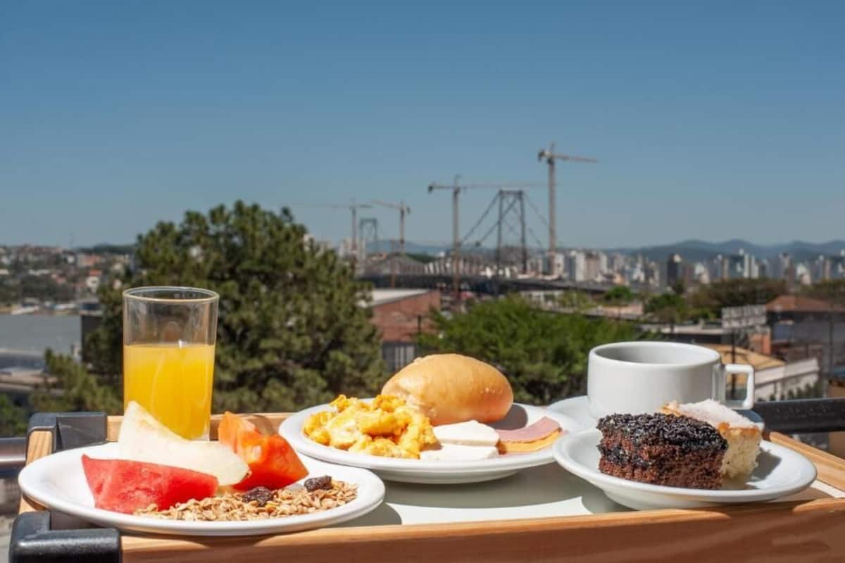A breakfast tray with various foods and a glass of orange juice, set against a scenic city view with cranes and buildings.