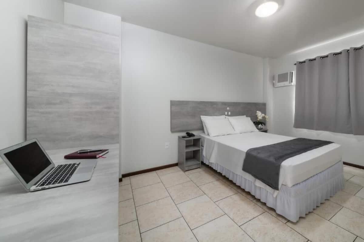 A modern hotel room with a bed, a desk with a laptop, a nightstand, and a bedside table with a lamp.