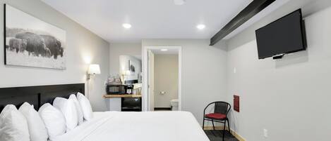 A hotel room with a large bed, a flat-screen TV mounted on the wall, a small kitchenette, and a chair.