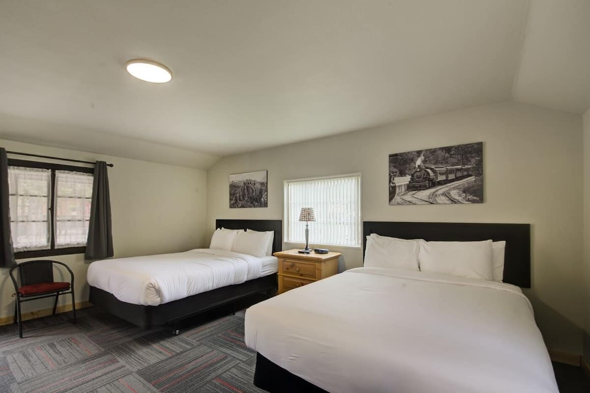 Una habitación de hotel con dos camas individuales, un pequeño escritorio con lámpara, una silla y dos ventanas con cortinas.