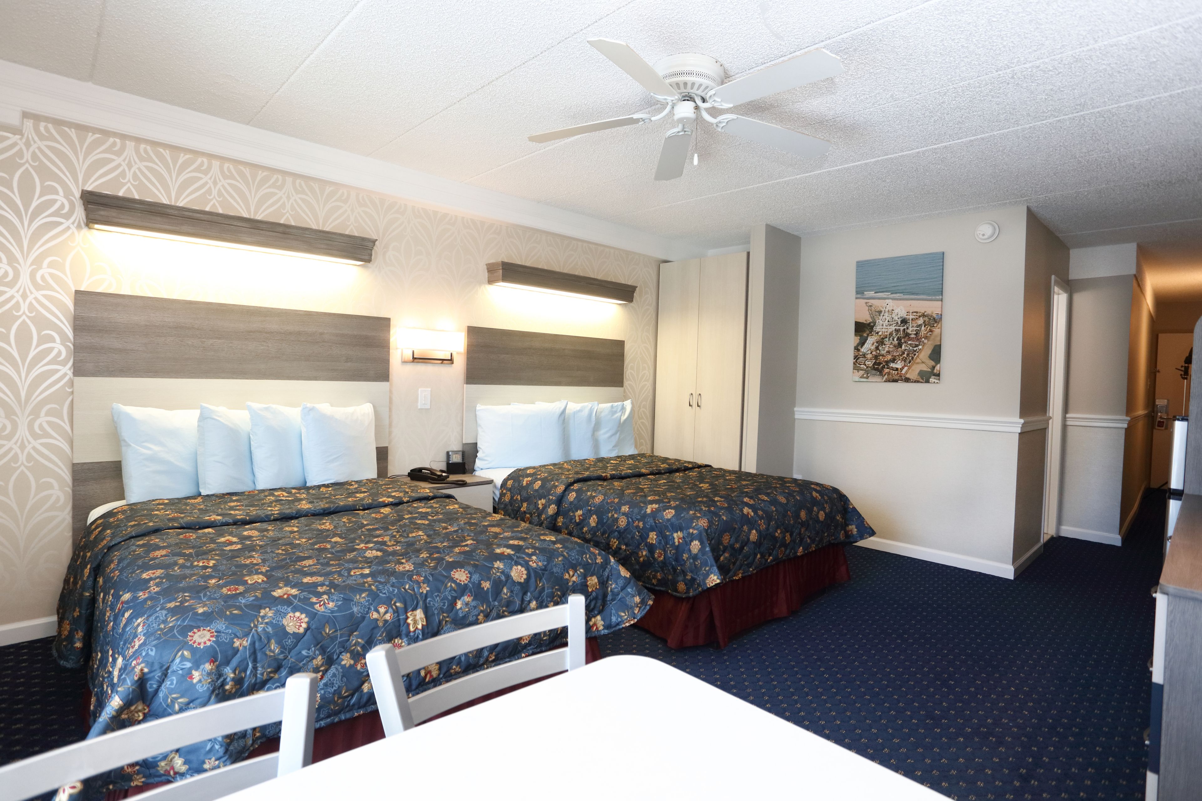 A hotel room with two beds, a desk, a ceiling fan, and a wall-mounted light.