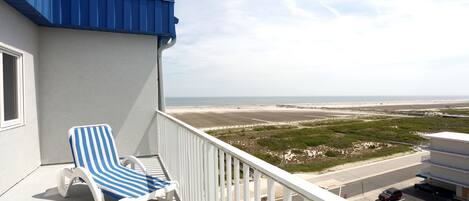 A balcony with a blue-striped lounge chair, a view of the beach, and a parking area.