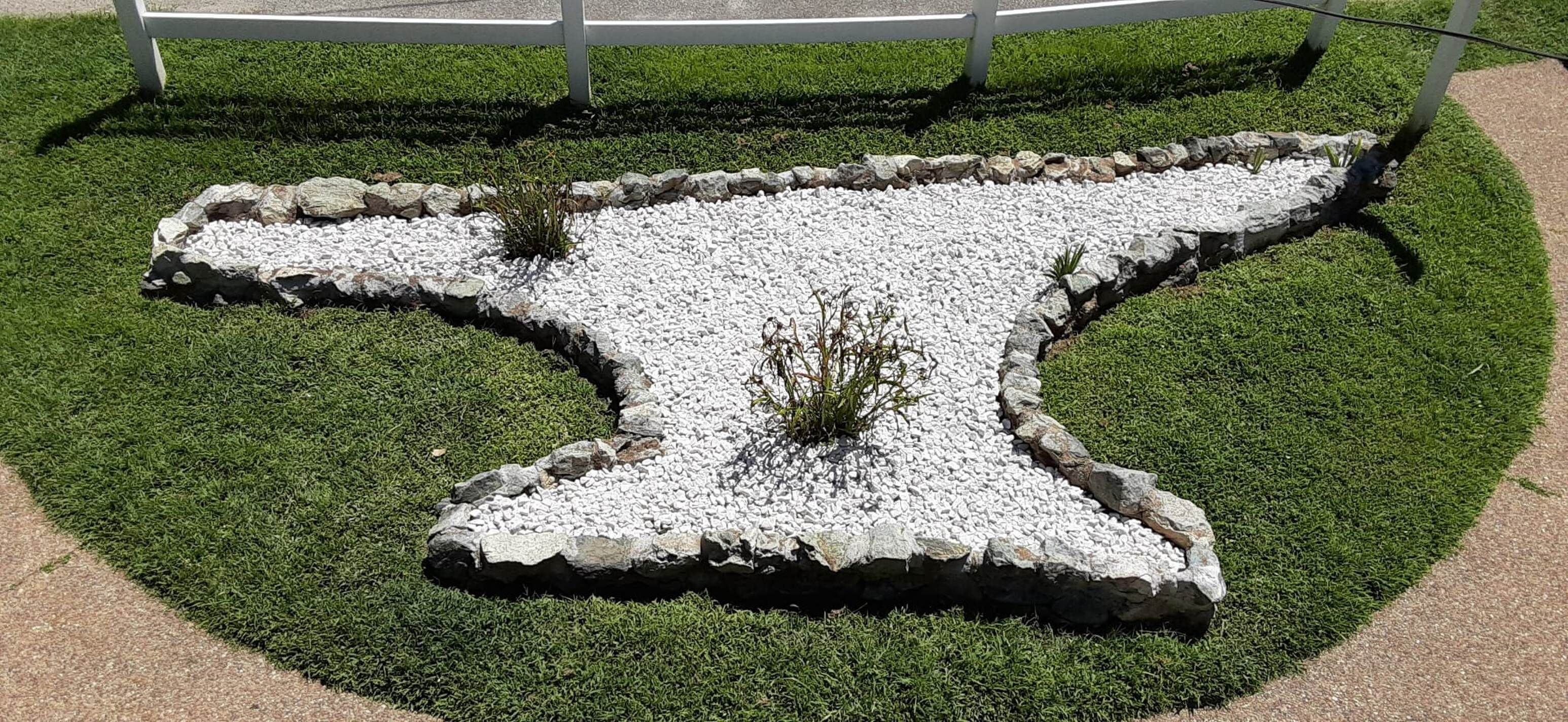 A cross-shaped garden bed with white pebbles and green grass, surrounded by a white fence.
