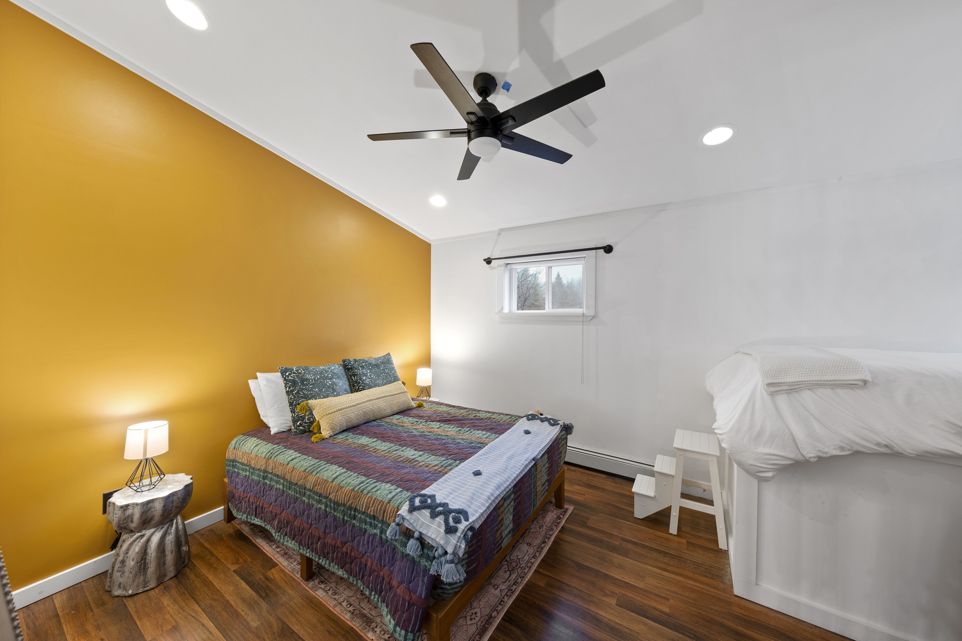 Un dormitorio con cama, ventilador de techo, mesita de noche, ventana y una silla blanca.