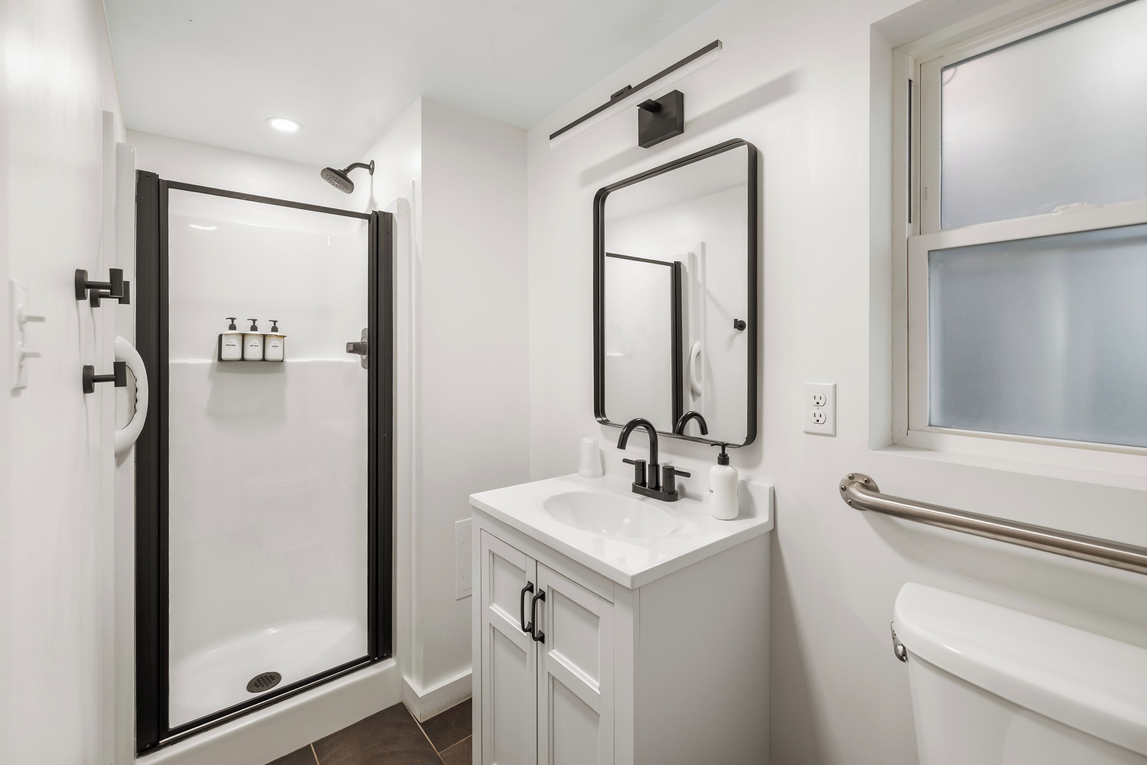 A modern bathroom with a glass-enclosed shower, a white sink with a mirror above it, and a toilet with a handrail.