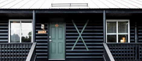 Una cabaña de troncos con una puerta verde, escalones que llevan a la entrada y una 'X' grande en la pared.