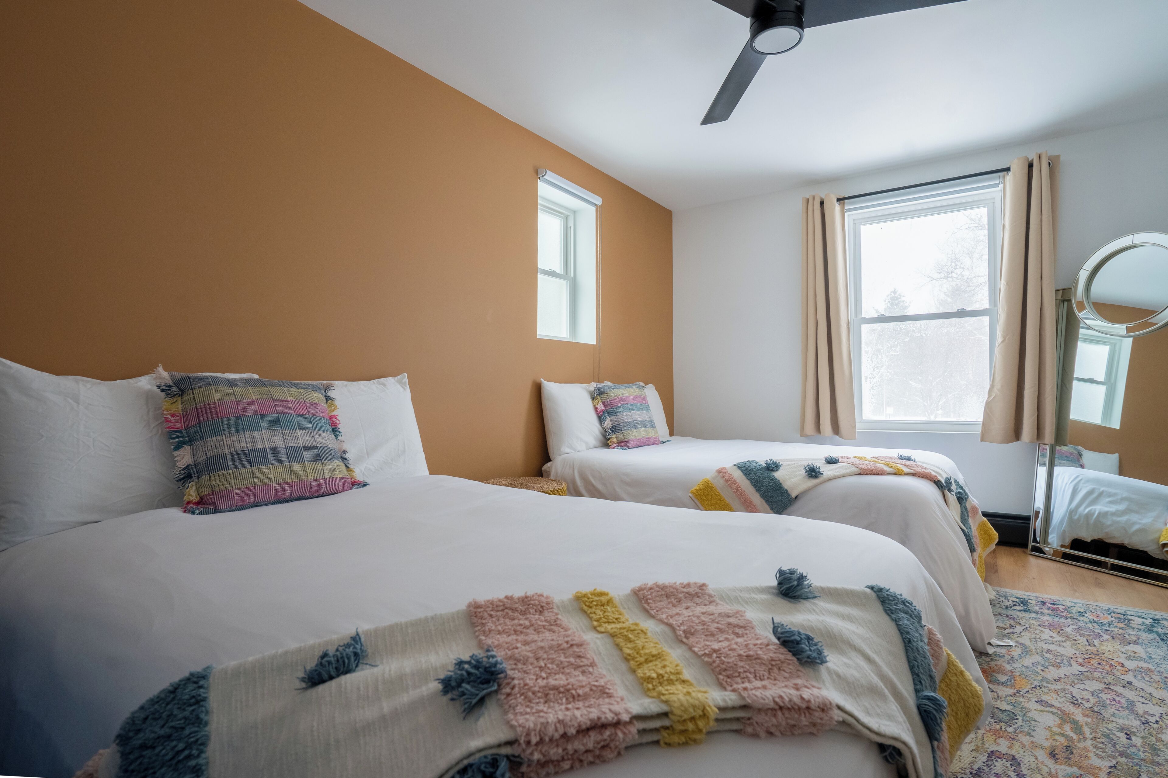 A hotel room with two beds, a ceiling fan, a window with curtains, and a mirror.