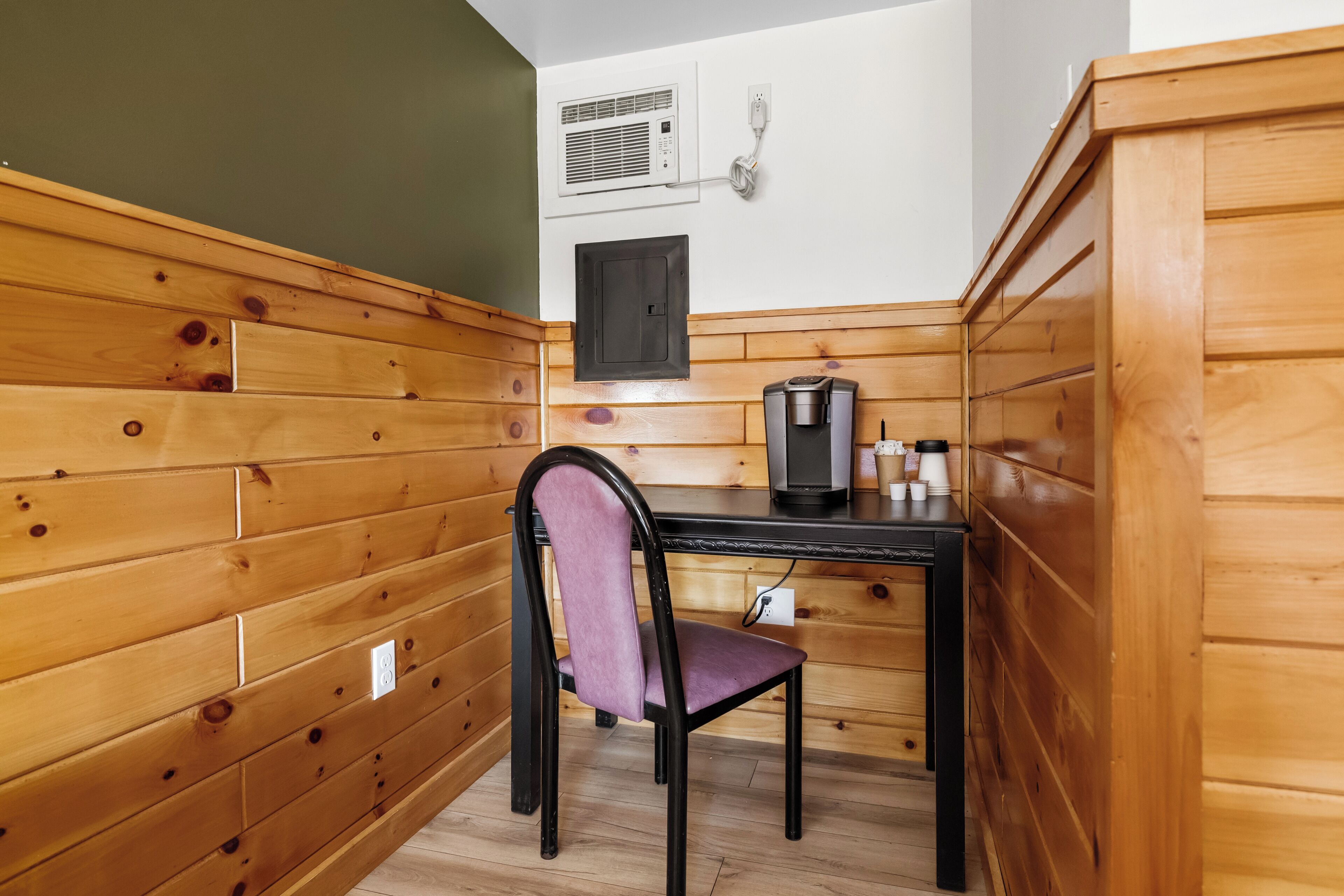 Une petite pièce aux murs en bois, avec un bureau noir, une chaise violette et une machine à café.
