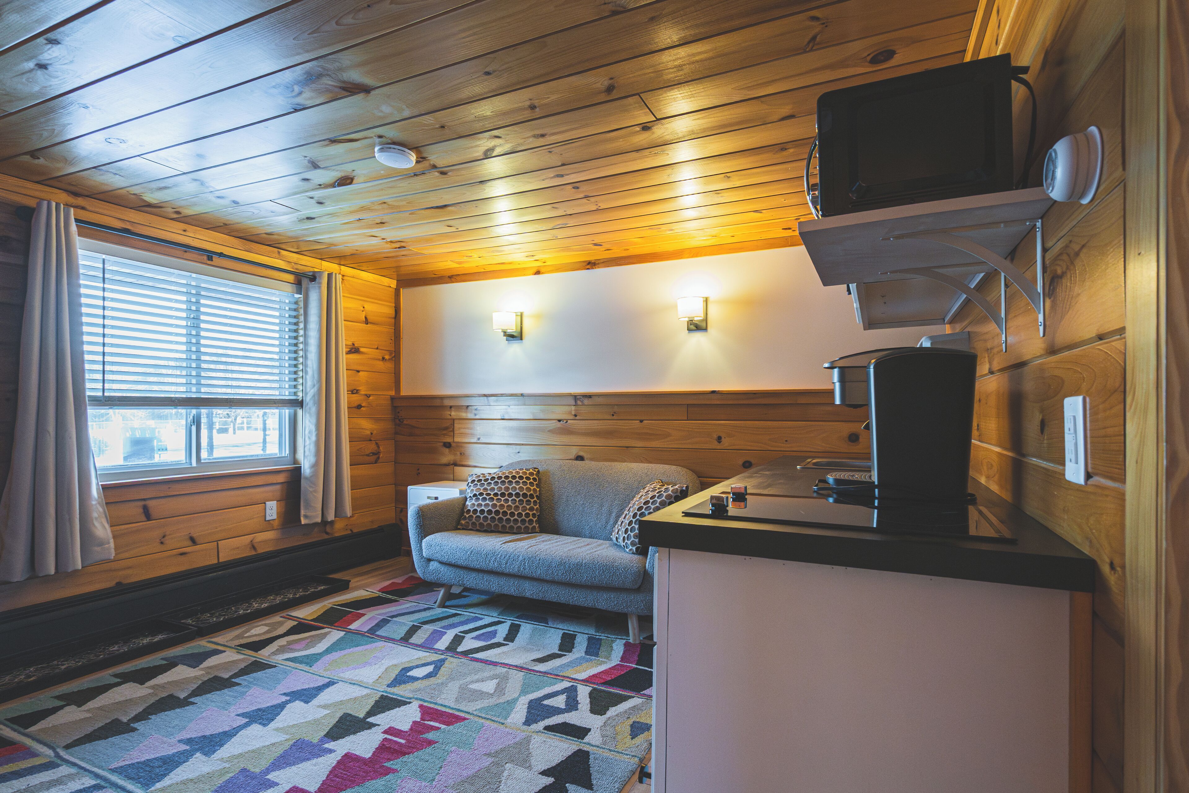 A cozy cabin interior with a wooden ceiling, a sofa with patterned cushions, a small kitchen area, and a window with curtains.