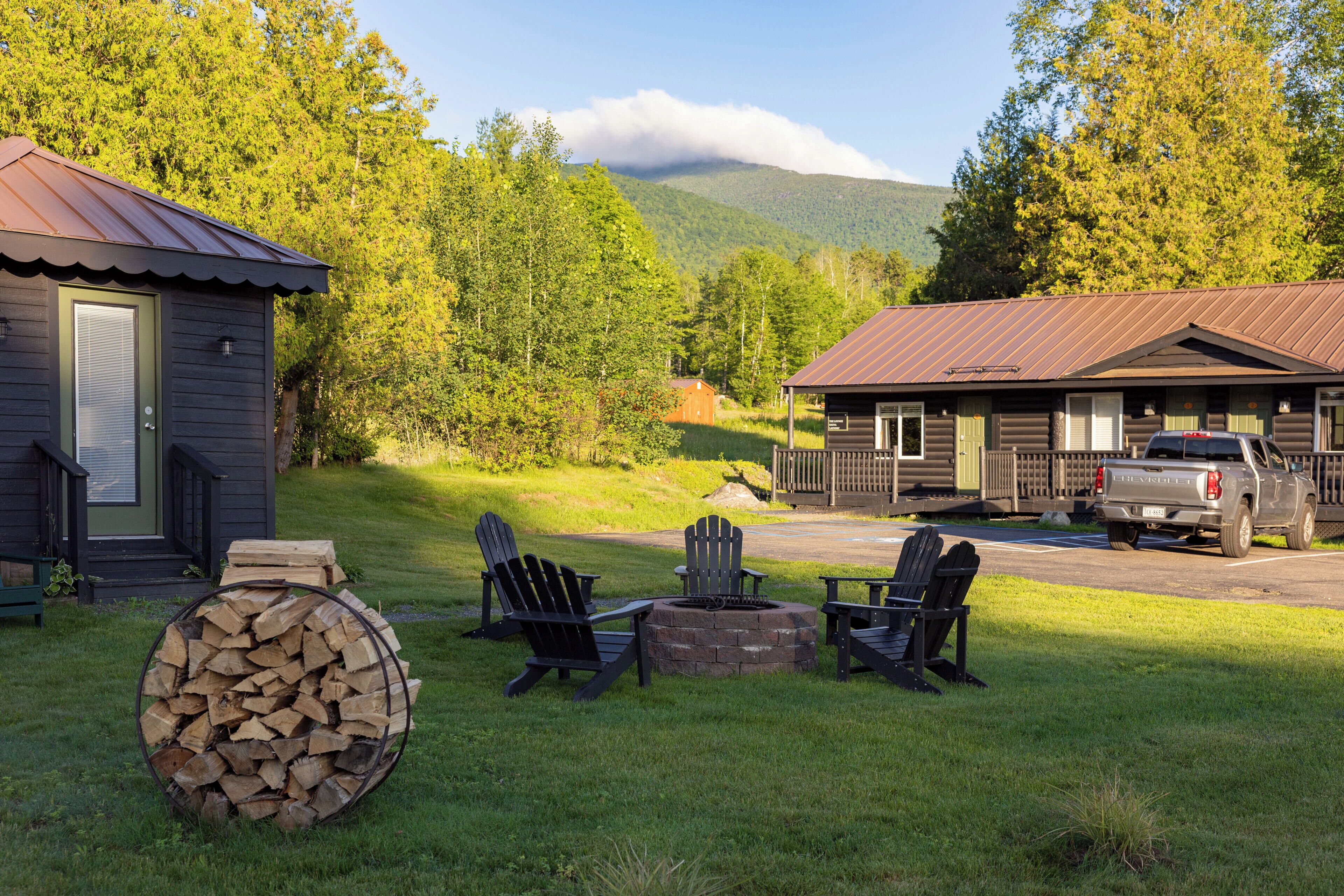 Eine Hütte mit Metall-Dach, Feuerstelle und Adirondack-Stühlen in einer Wiese mit Bäumen und Bergen im Hintergrund.