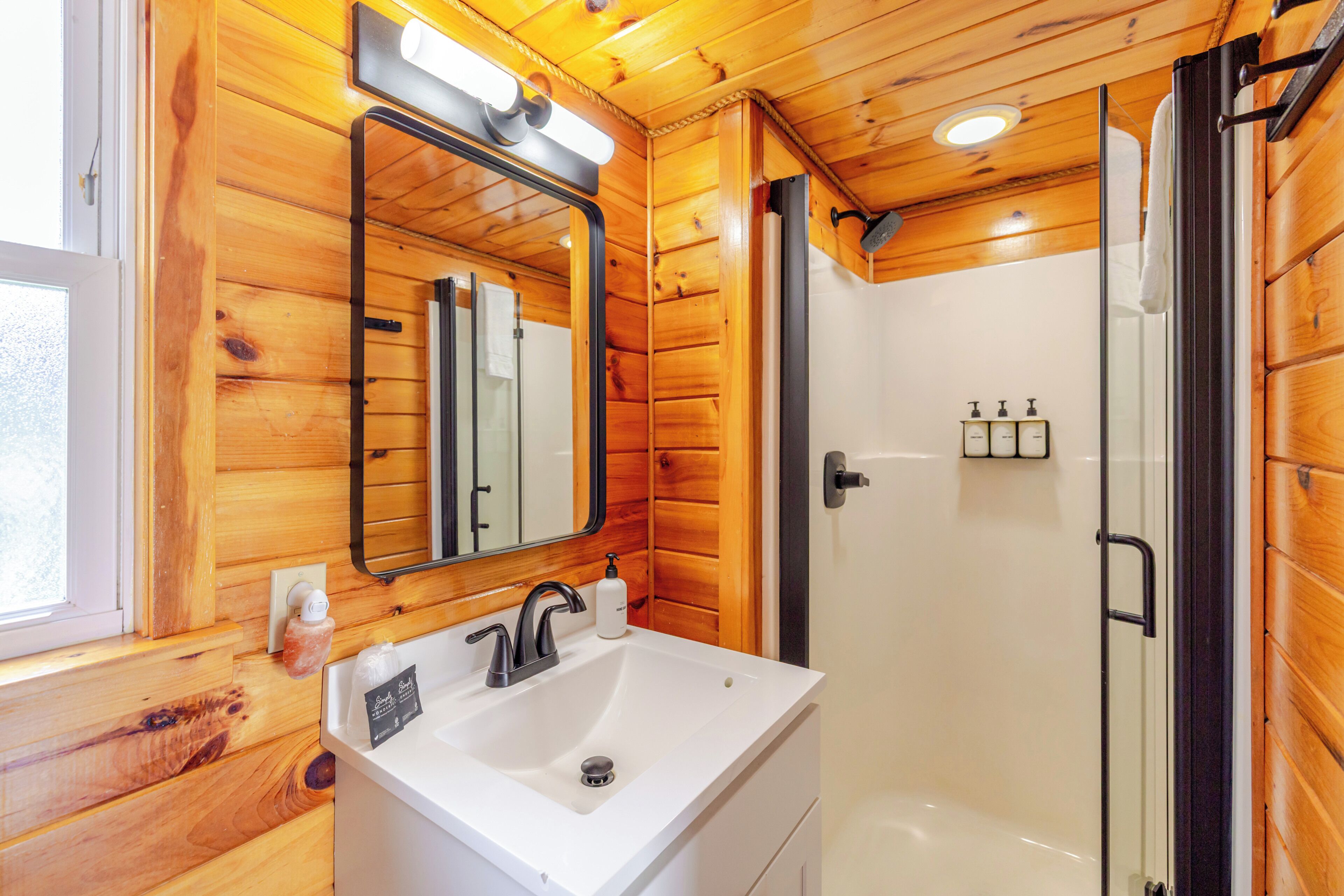 Baño de cabaña de madera con lavamanos, espejo y ducha blancos.