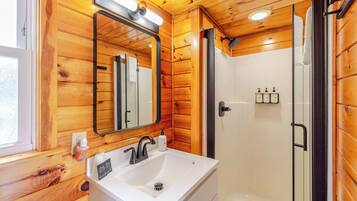 Baño de cabaña de madera con lavamanos, espejo y ducha blancos.