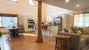 Ein geräumiger Wohnbereich mit einem Holzesstisch, Stühlen, einer Couch und einem kleinen Holzschemel. Es gibt eine Küchenzeile mit Schränken und Geräten sowie ein großes Fenster mit Vorhängen.
