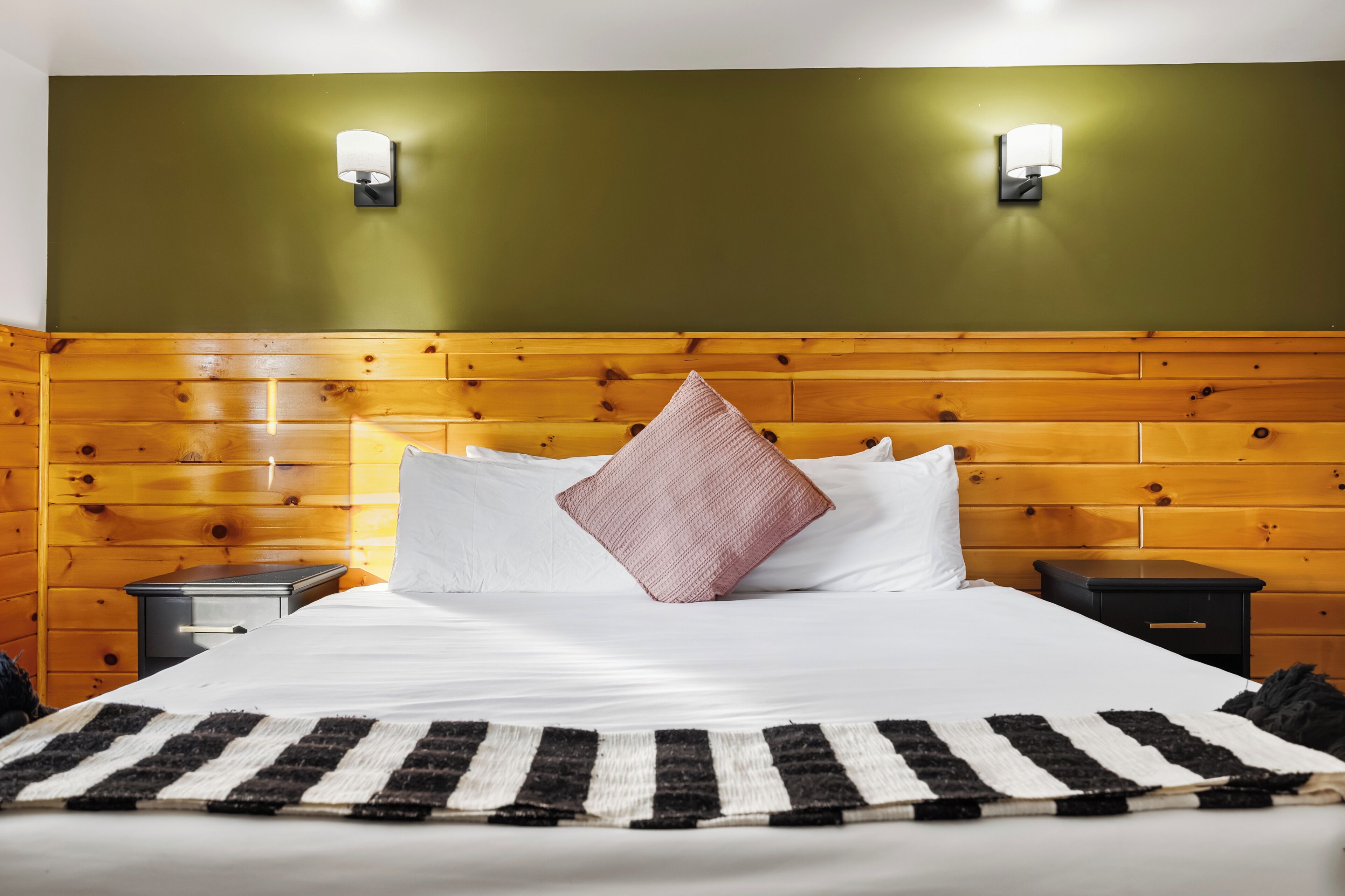 A hotel room with a wooden headboard, two bedside tables, a bed with white bedding, a striped rug, and two wall-mounted light fixtures.