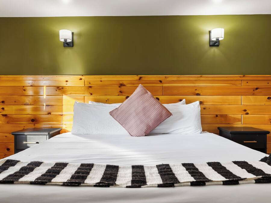 A hotel room with a wooden headboard, two bedside tables, a bed with white bedding, a striped rug, and two wall-mounted light fixtures.