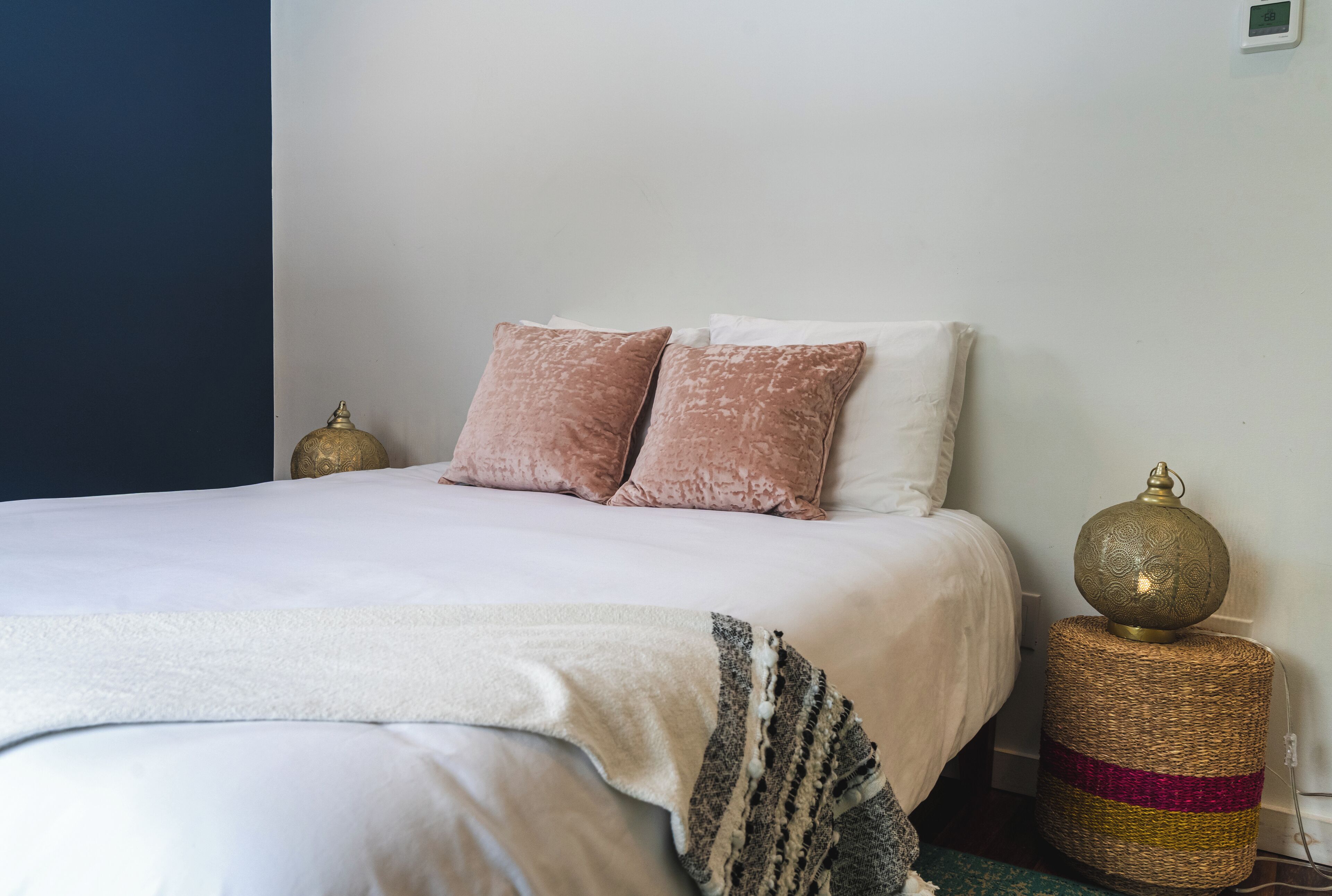A neatly made bed with white bedding and two pink pillows, a decorative woven basket with a golden ornament, and a wall-mounted thermostat.