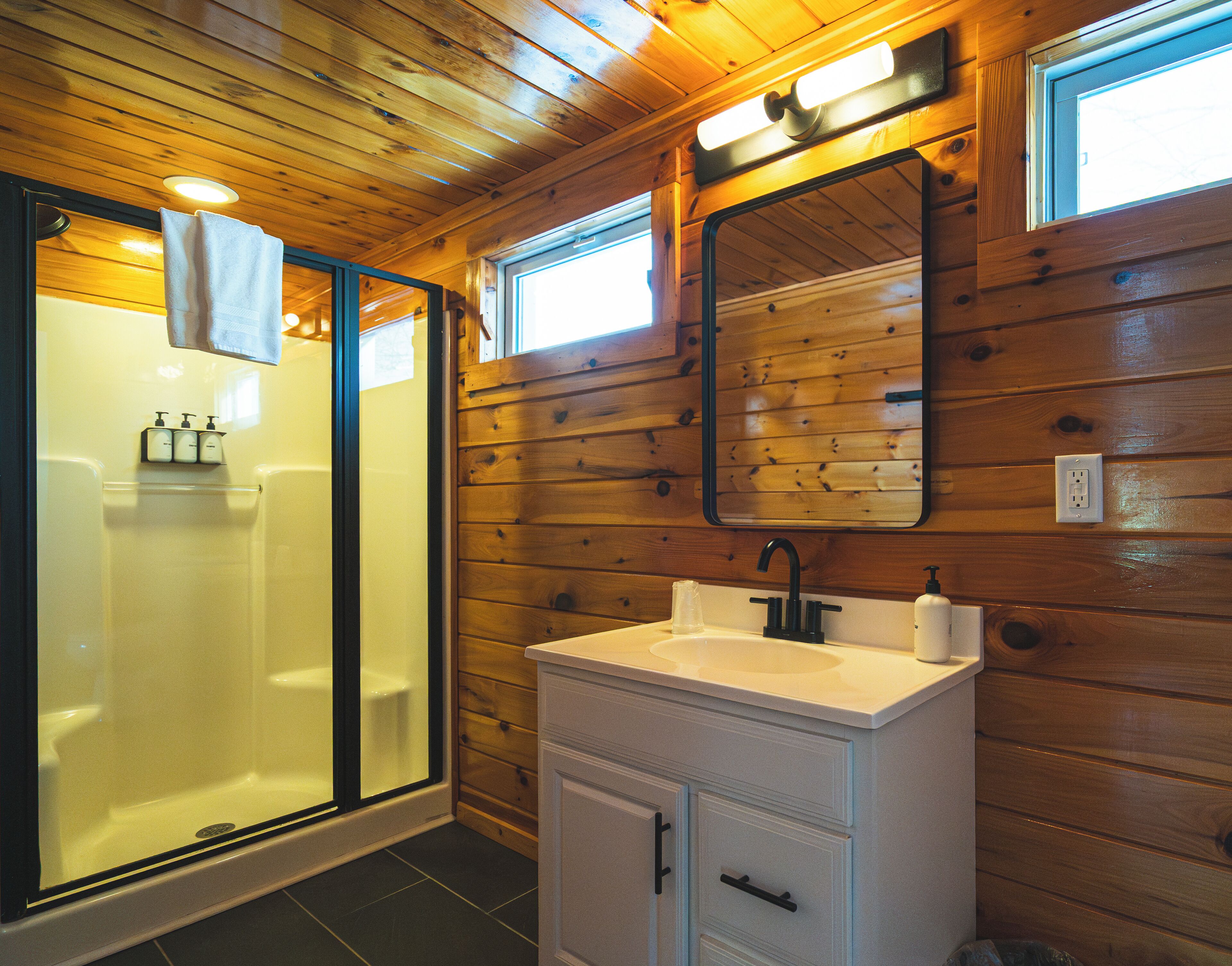 A wooden-paneled bathroom with a glass-enclosed shower, a white sink with a mirror above it, and a small window.