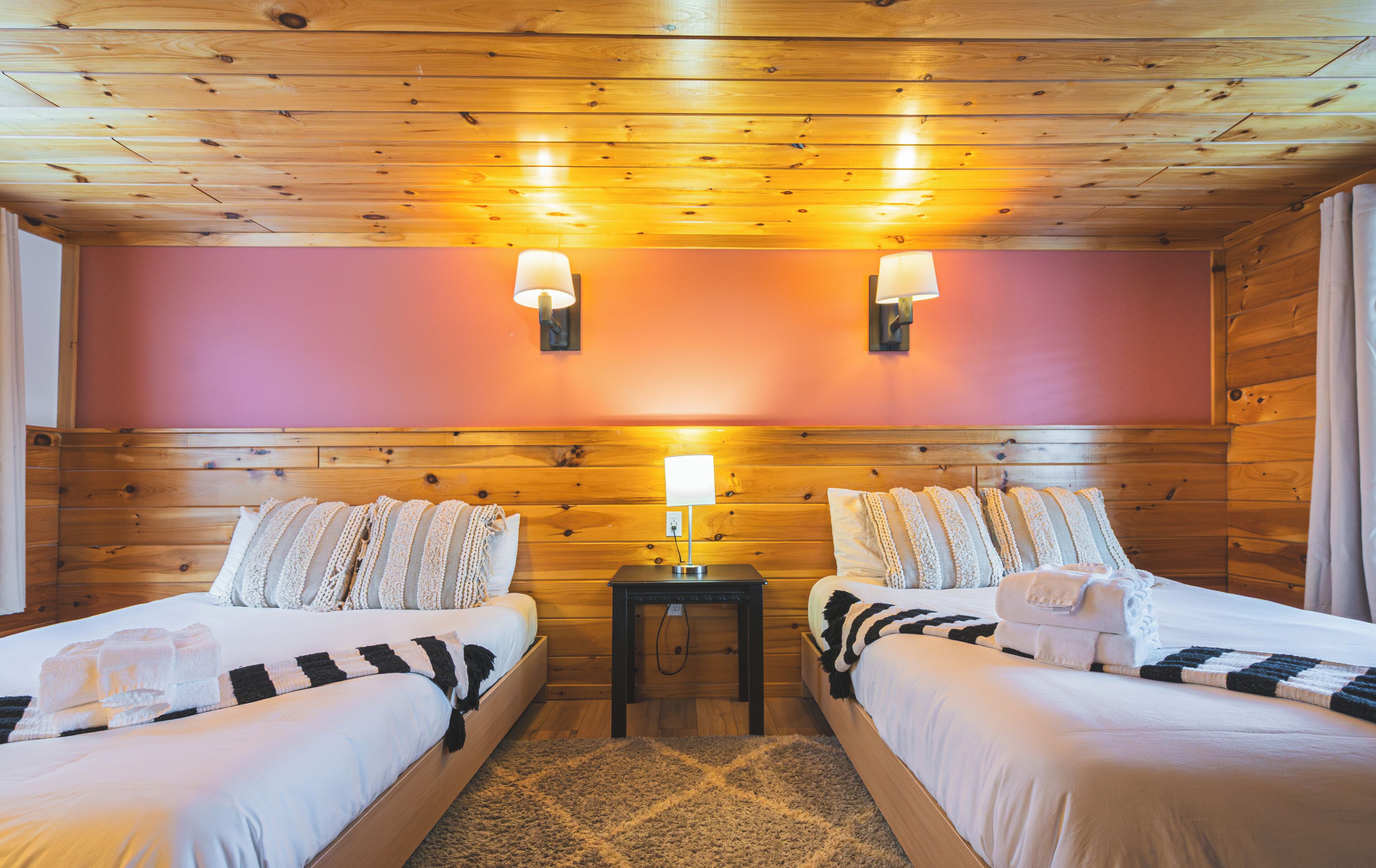 A room with two beds, wooden ceiling, and a small table with a lamp.