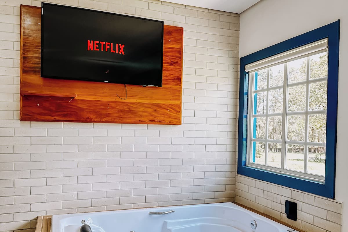 A modern bathroom with a wooden bathtub, a large mirror, and a wall-mounted TV displaying the Netflix logo.