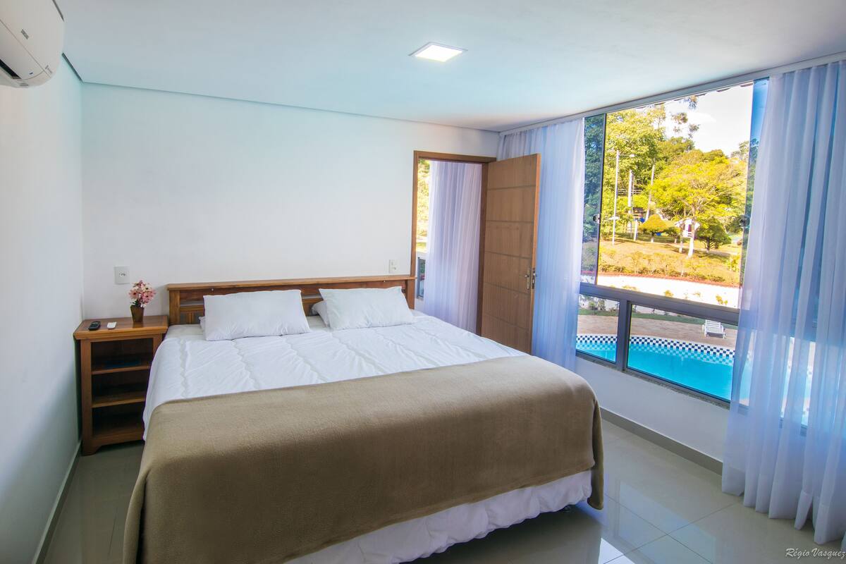 A bedroom with a large bed, a nightstand, a window with a view of a swimming pool and trees.