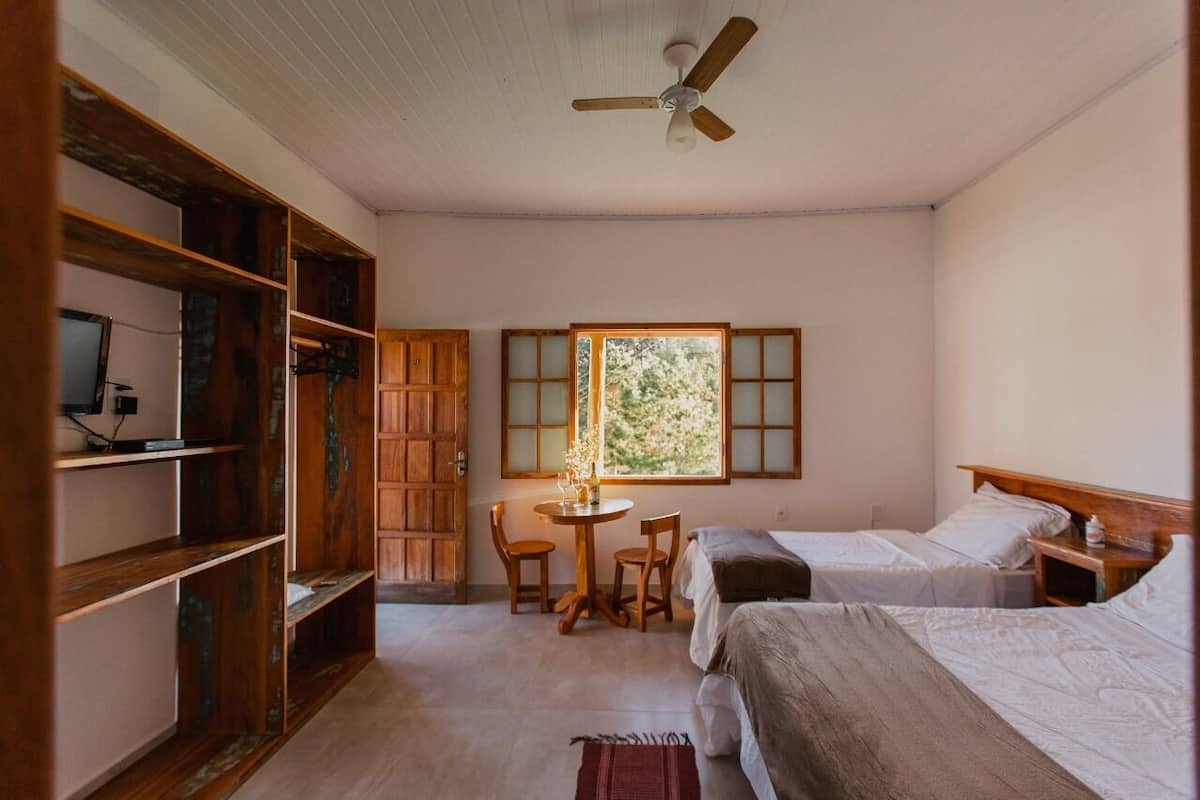 A room with two beds, a wooden bookshelf, a desk with chairs, a window with a view, and a ceiling fan.