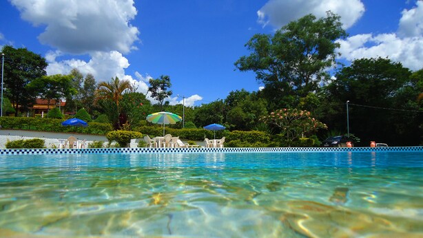 A clear swimming pool with lounge chairs and umbrellas, surrounded by lush greenery and trees. - Hotel Pontal de Tiradentes (Tiradentes)