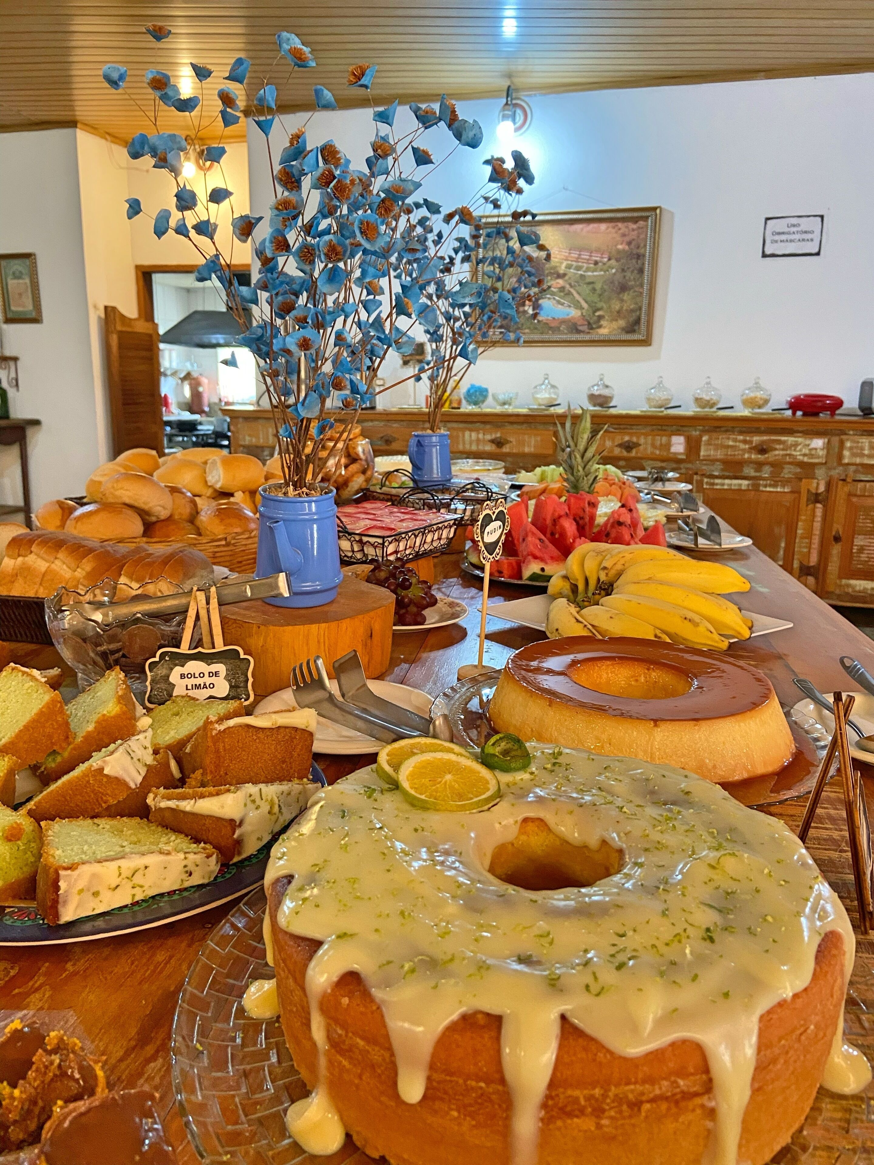 Una mesa de buffet con diversos alimentos, incluyendo un pastel bundt grande con cobertura blanca, una bandeja de frutas con plátanos y sandía, y varios tipos de pan.