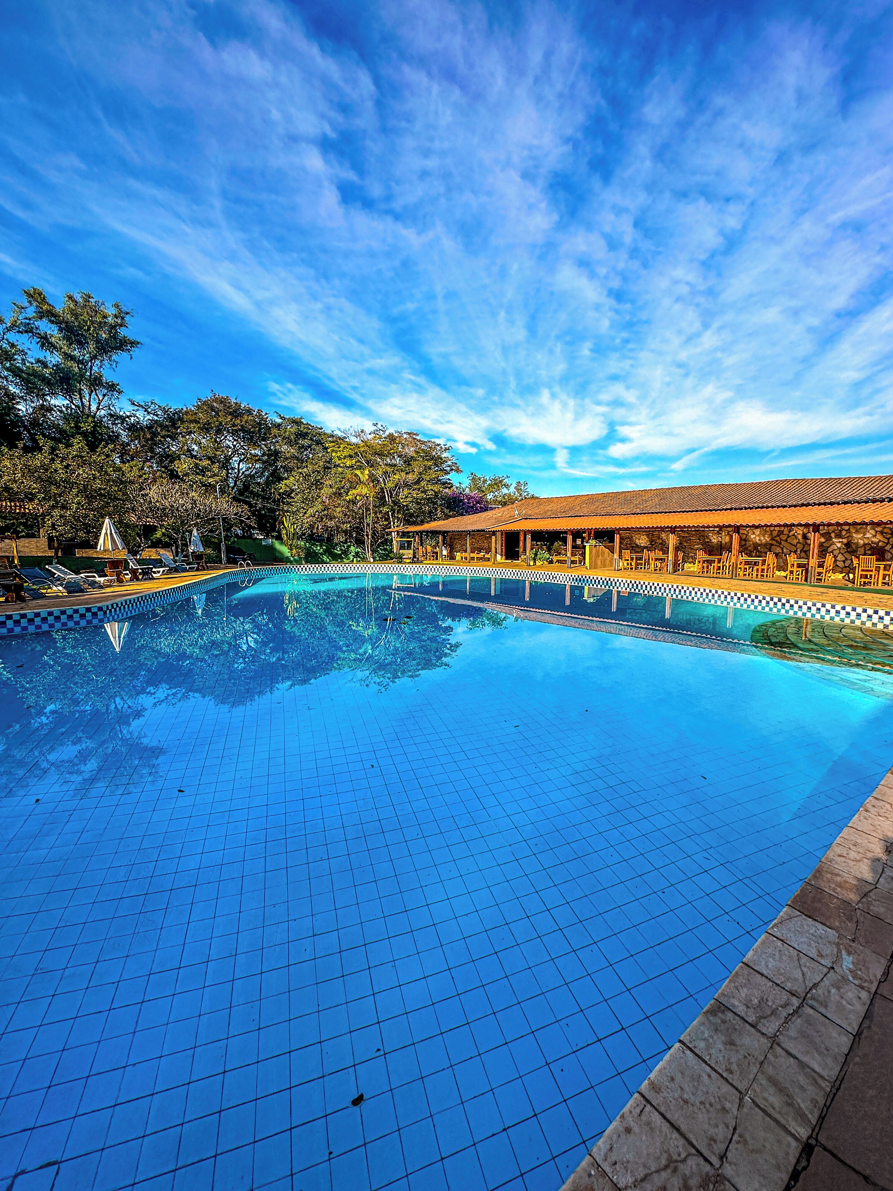 A swimming pool with tiled flooring, surrounded by lounge chairs and umbrellas, with a building in the background.